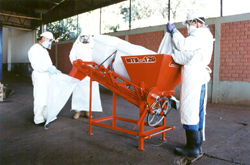 Três manipuladores vestidos com EPIs (aventais, luvas, mascaras e gorros) ao redor de maquinário destinado a tratamento de sementes "on farm".